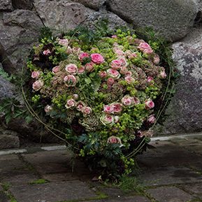 Trauerfloristik Herzförmiger Blumenarrangement mit rosa und grünen Blüten vor einer steinernen Wand.